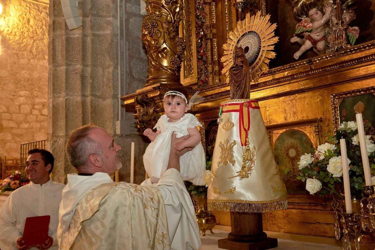 Sacerdote sosteniendo a un niño ante un altar religioso dorado con velas y estatuas.