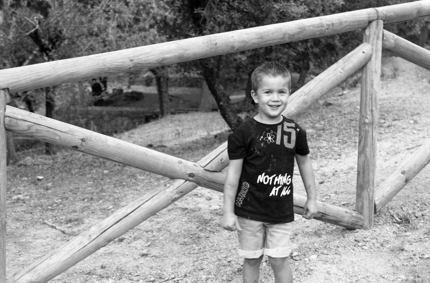 Un niño pequeño sonríe junto a una valla de madera en un camino de tierra, vestido con una camiseta oscura y pantalones cortos.