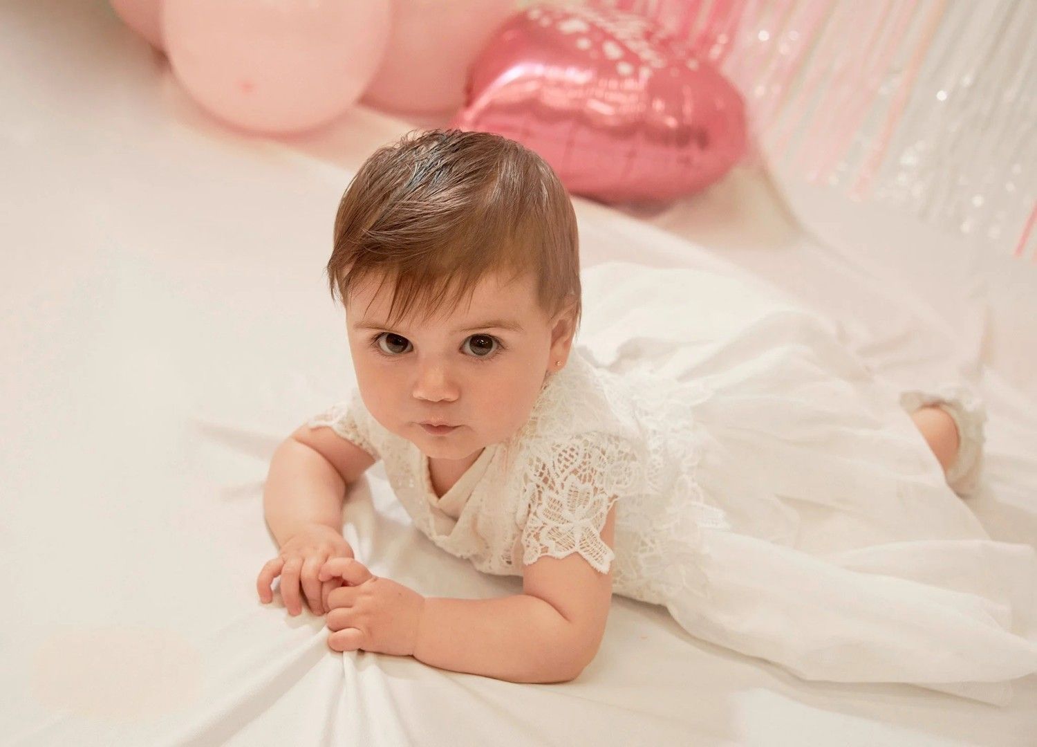 Bebé tumbado sobre una suave manta blanca, con un vestido blanco y globos de colores pastel de fondo.