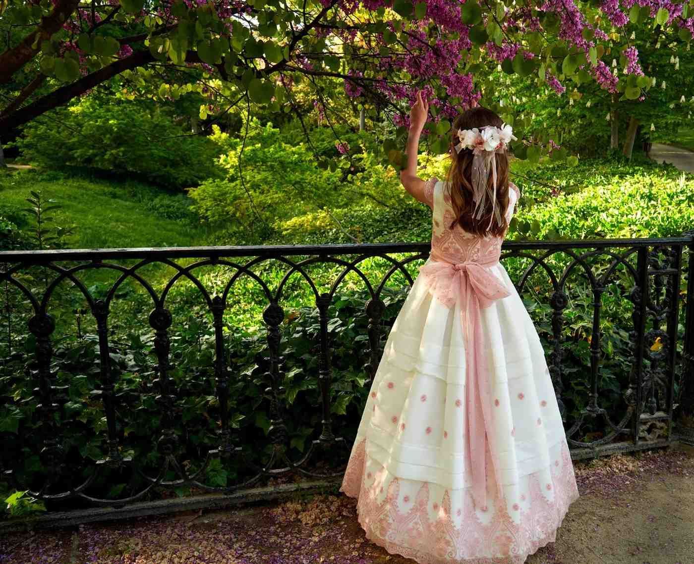 Una persona con un vestido rosa se estira junto a una valla negra bajo flores moradas en plena floración en un jardín.