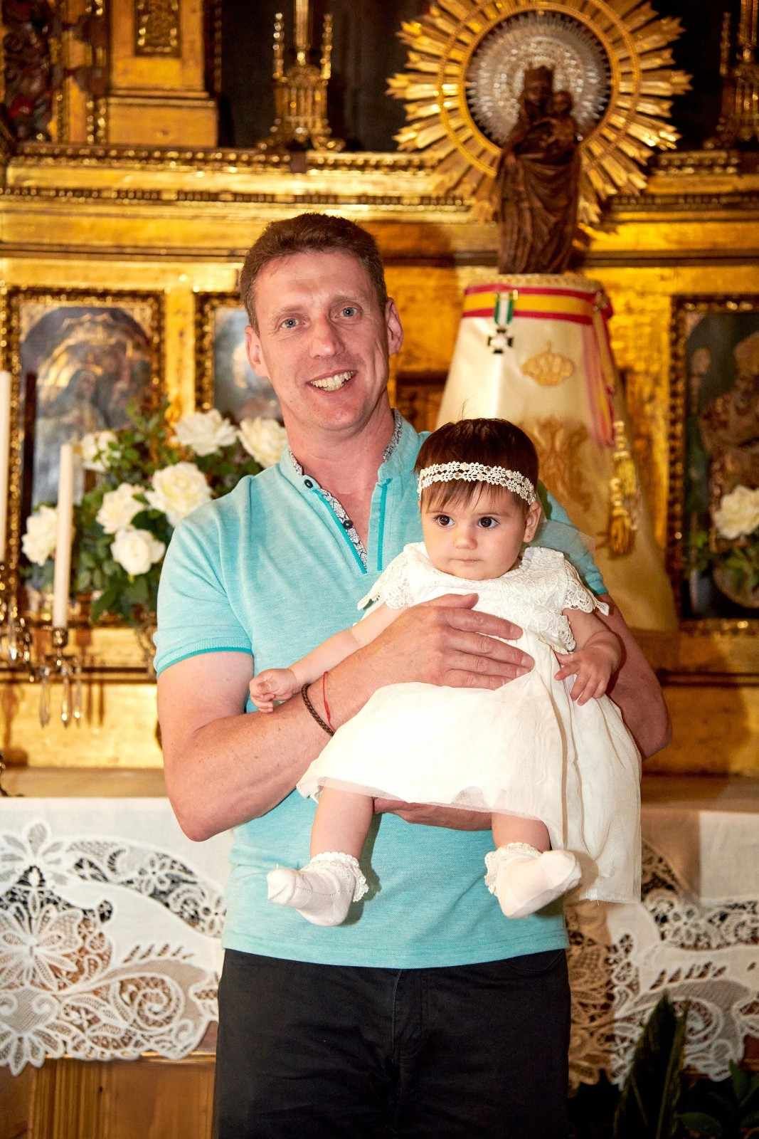 Un hombre sostiene a un bebé vestido de blanco dentro del área del altar de una iglesia decorada con oro.
