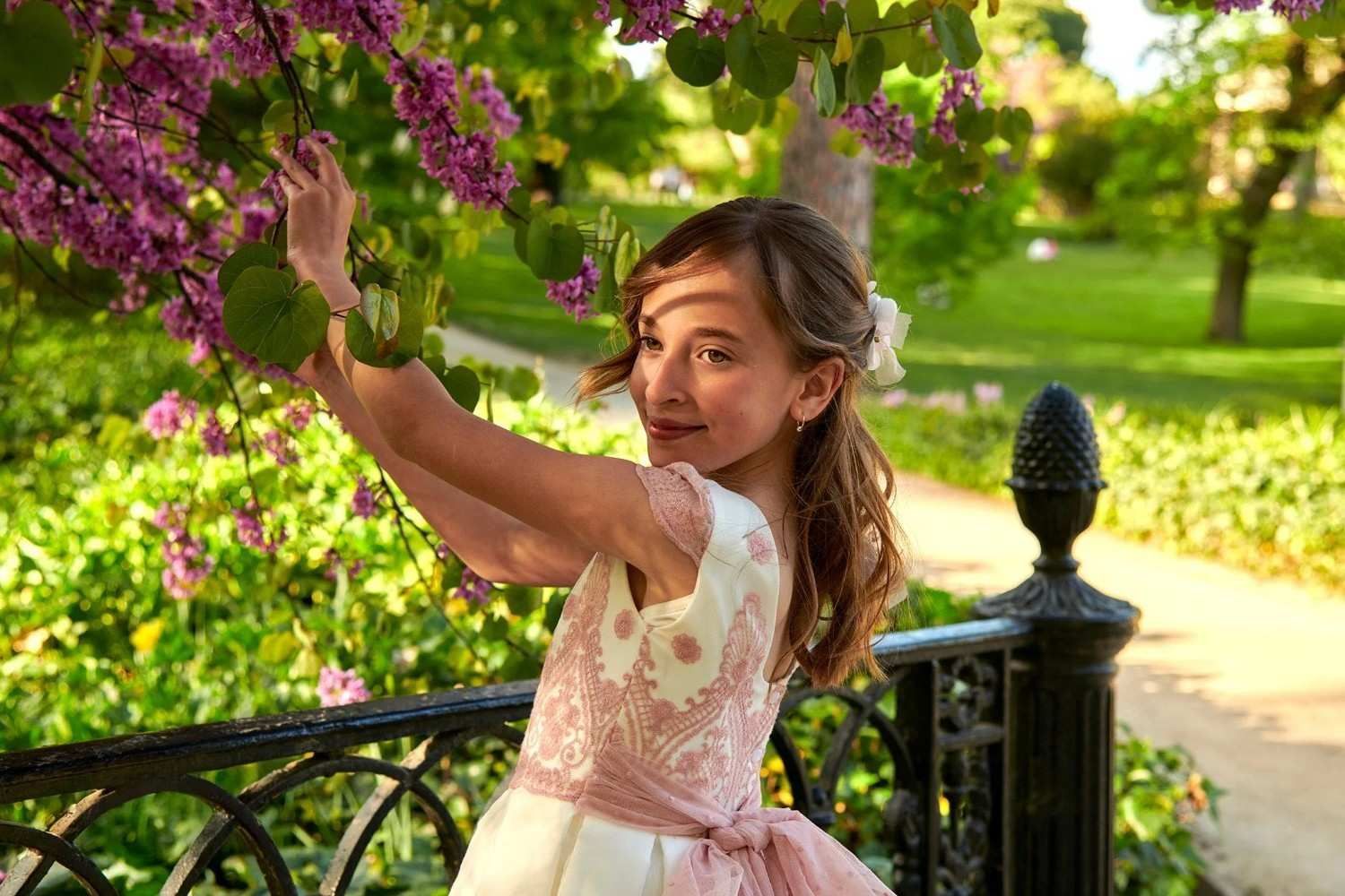 Una niña con un vestido rosa intenta alcanzar unas flores moradas en un jardín soleado.