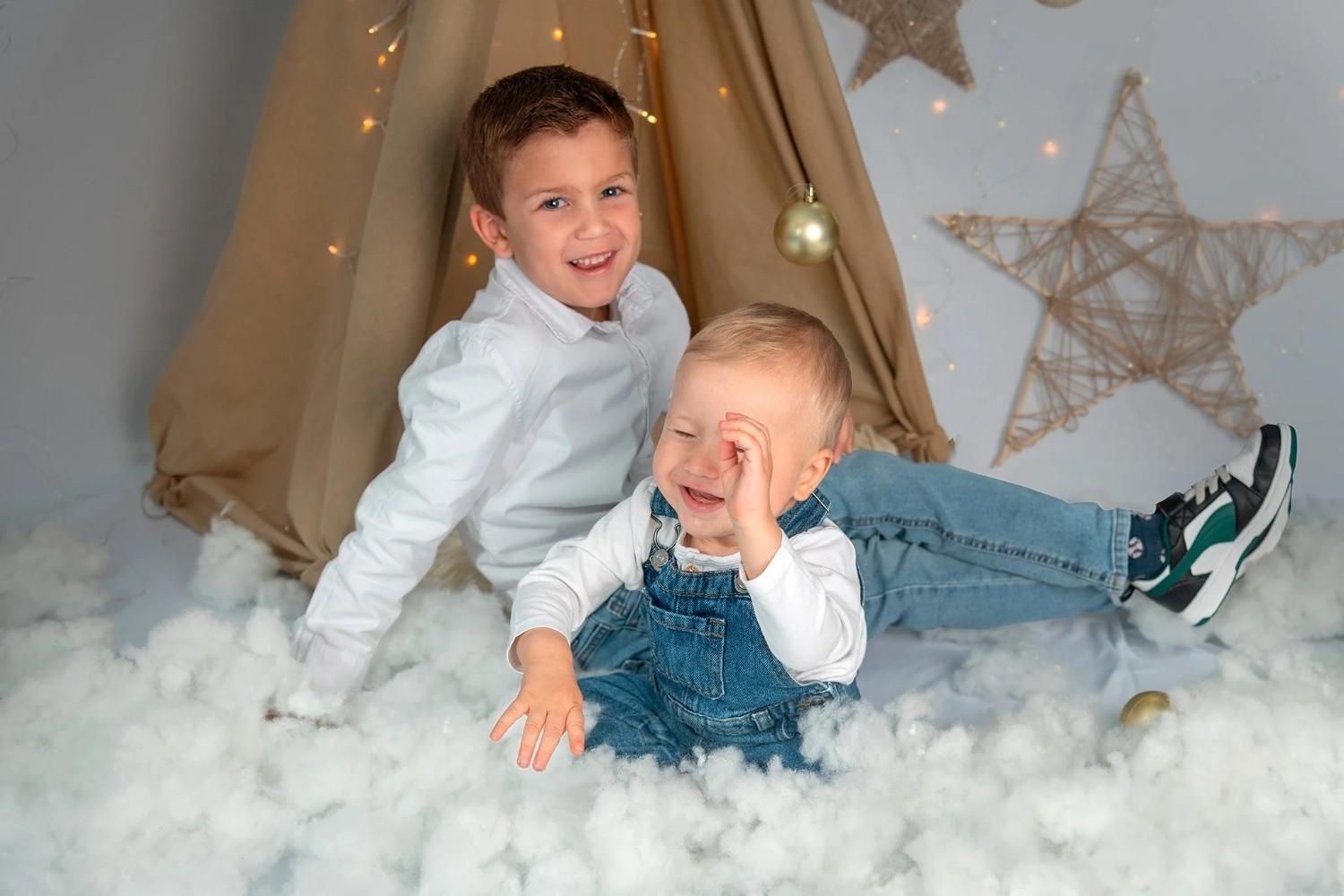 Dos niños con camisas blancas y overoles de mezclilla juegan en nieve artificial blanca y esponjosa, sonriendo en una acogedora escena de tienda de campaña.