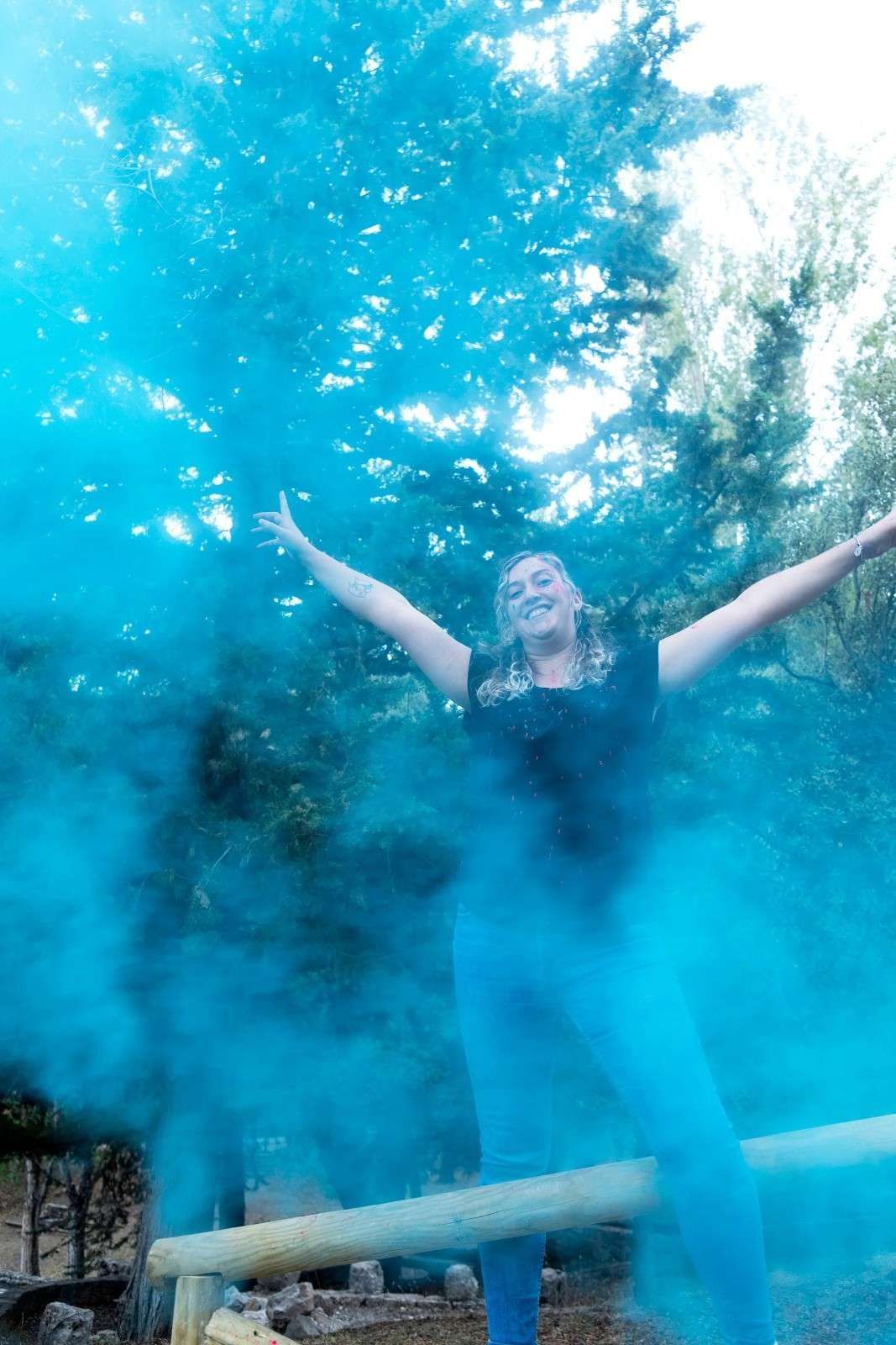 Una persona permanece de pie con los brazos en alto, rodeada de una nube de humo azul, al aire libre, cerca de una barandilla de madera.