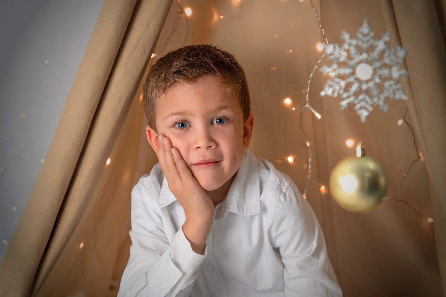 Un niño con una camisa blanca apoya la cara en una mano junto a adornos navideños y un adorno colgante.