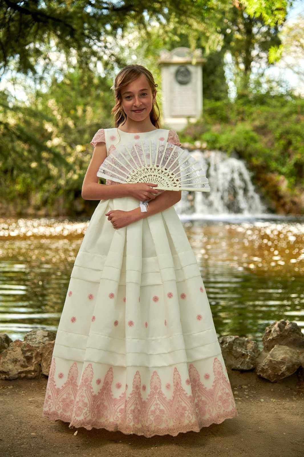 Mujer joven con un vestido blanco bordado sosteniendo un abanico de encaje junto a una cascada y un estanque.