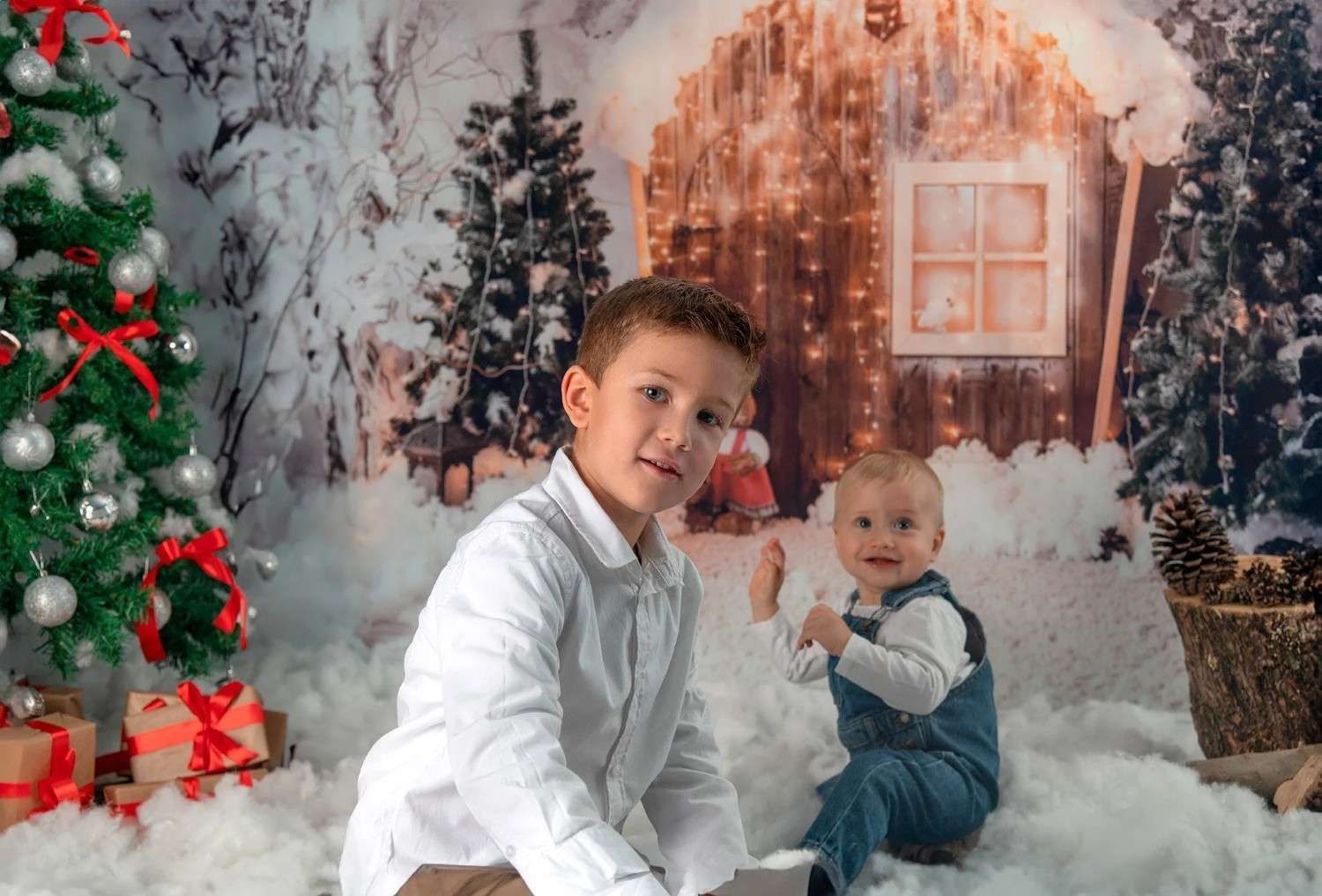 Dos niños vestidos de blanco posan en una escena navideña nevada con un árbol decorado y una cabaña iluminada de fondo.