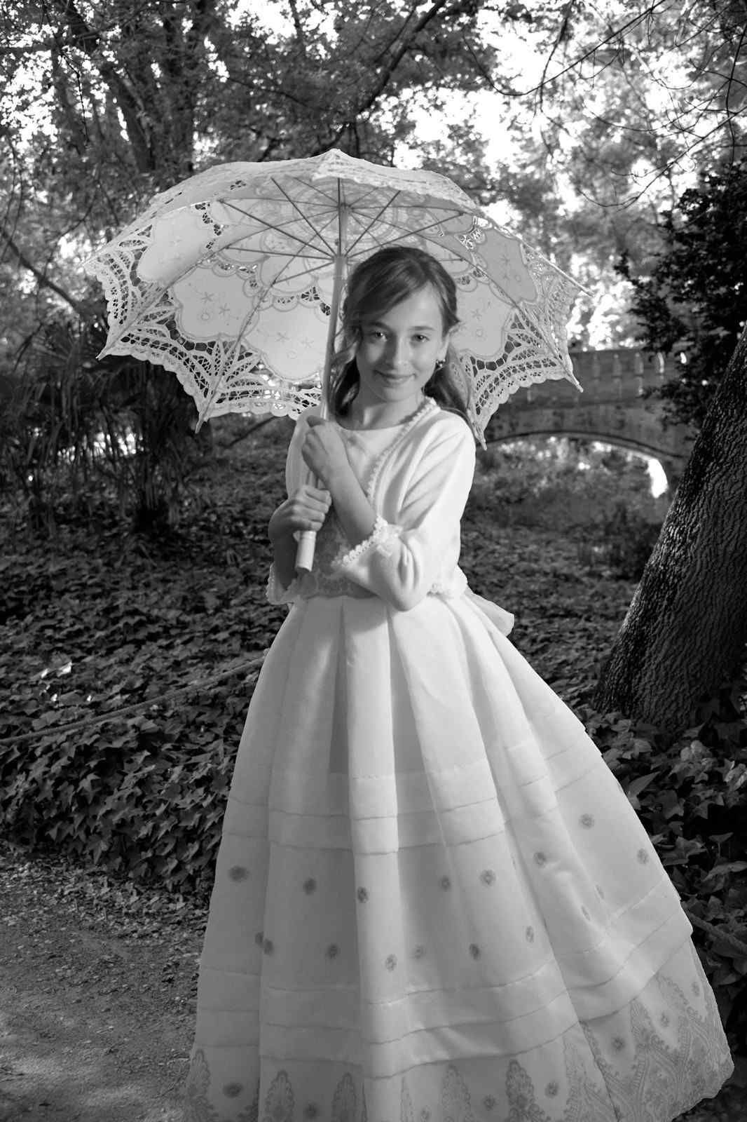Niña con un vestido blanco largo sosteniendo una sombrilla de encaje al aire libre bajo los árboles.