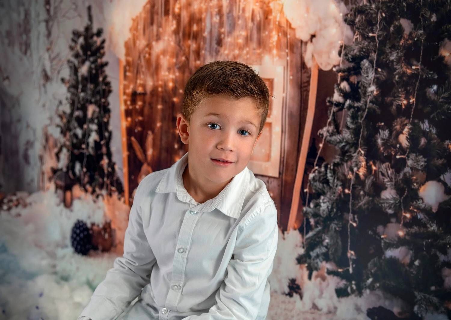 Un niño pequeño con una camisa blanca posa frente a un paisaje invernal nevado con luces cálidas.