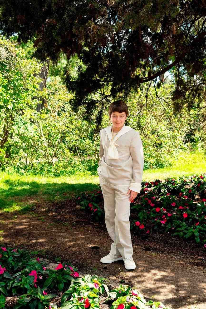 Una persona vestida con un traje blanco se encuentra de pie en un jardín soleado junto a flores rosas y vegetación.