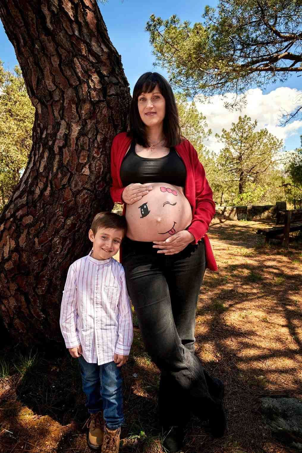 Una mujer embarazada con un cárdigan rojo está de pie con su hijo junto a un árbol en un parque soleado.