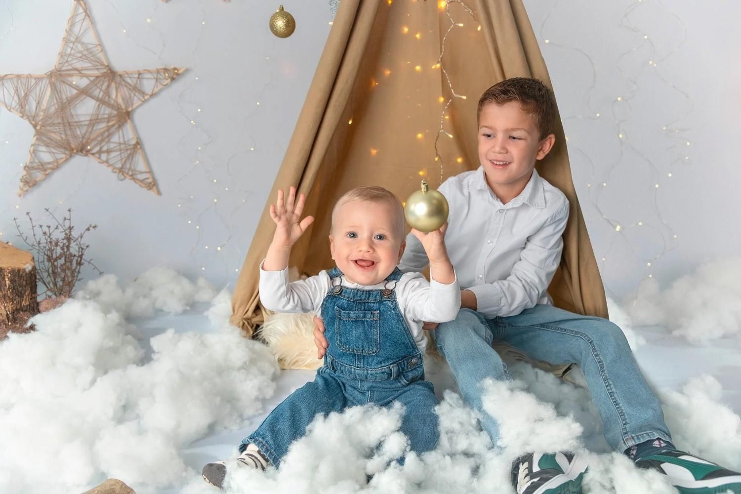 Dos niños vestidos de mezclilla están sentados sobre nubes esponjosas en un acogedor tipi con adornos dorados y luces cálidas.