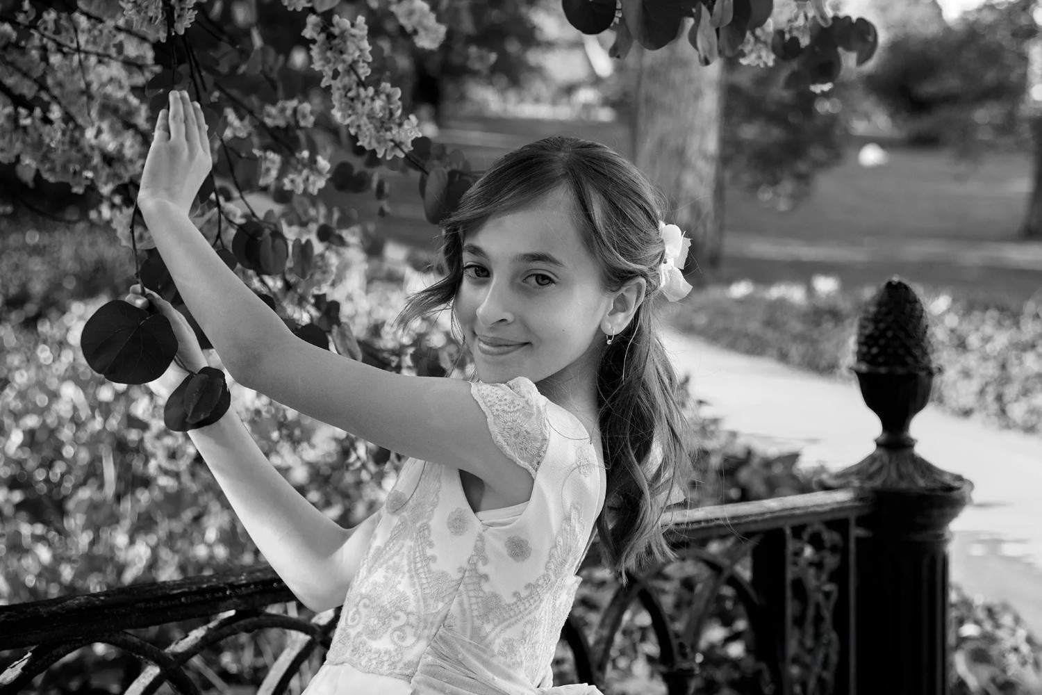 Una joven con un vestido blanco posa en un banco de jardín junto a una valla de hierro forjado.