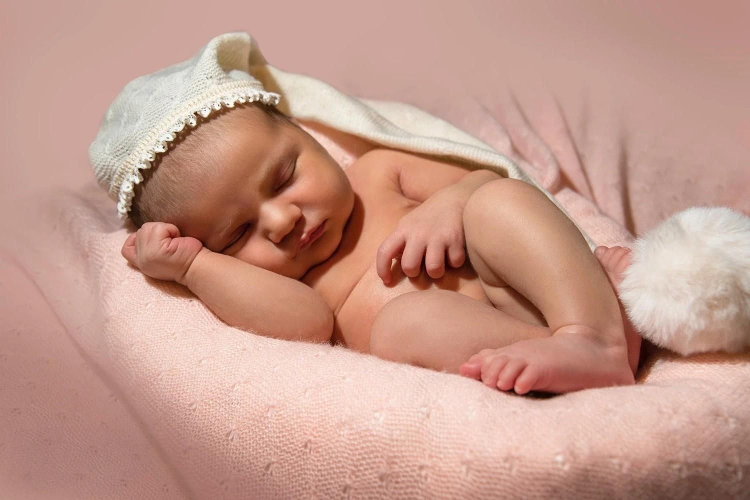 Bebé dormido acurrucado en una manta rosa, con un gorrito de encaje blanco.