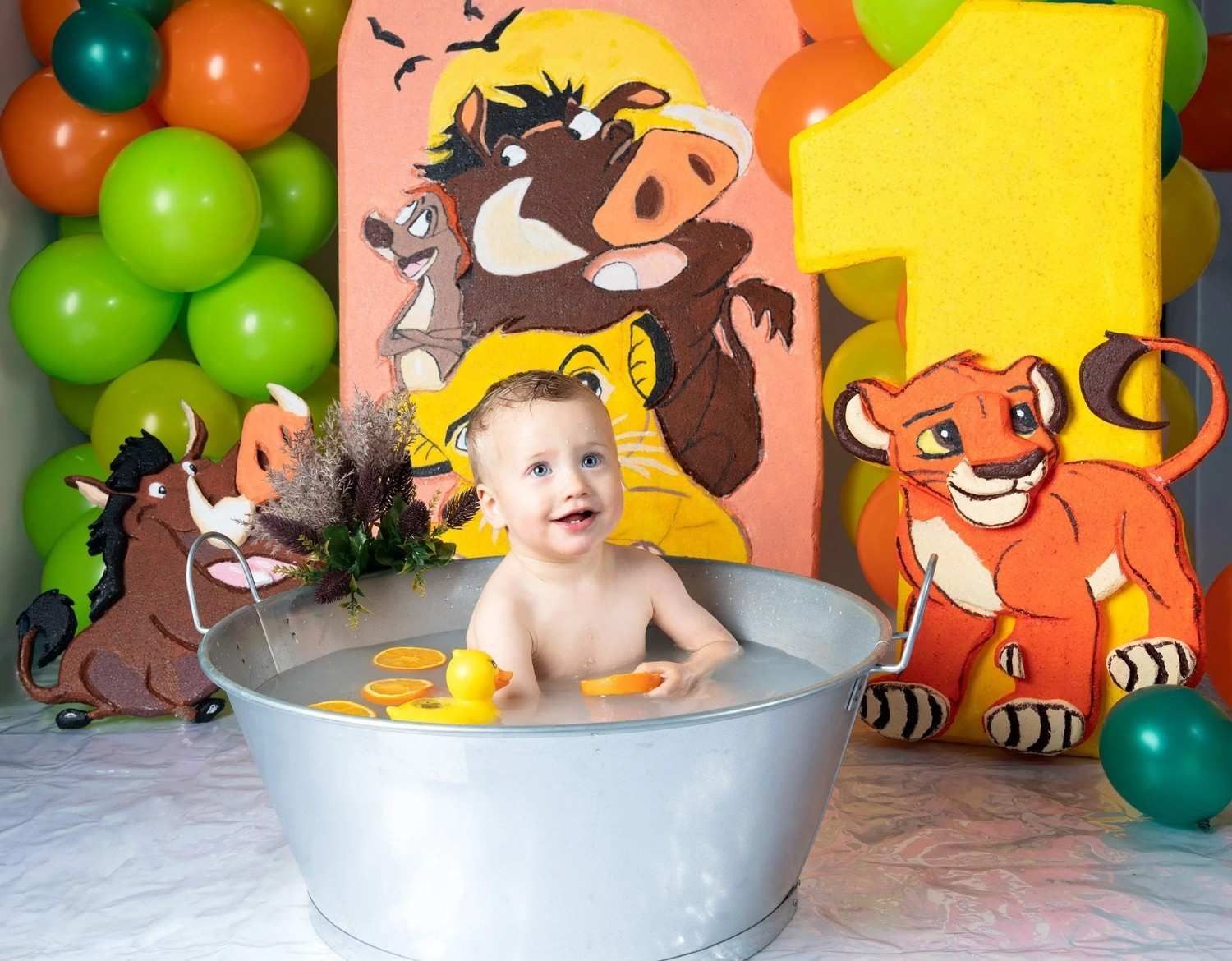 Bebé en una bañera plateada con patitos amarillos, frente a coloridas decoraciones con temática de leones y un 1 amarillo gigante.