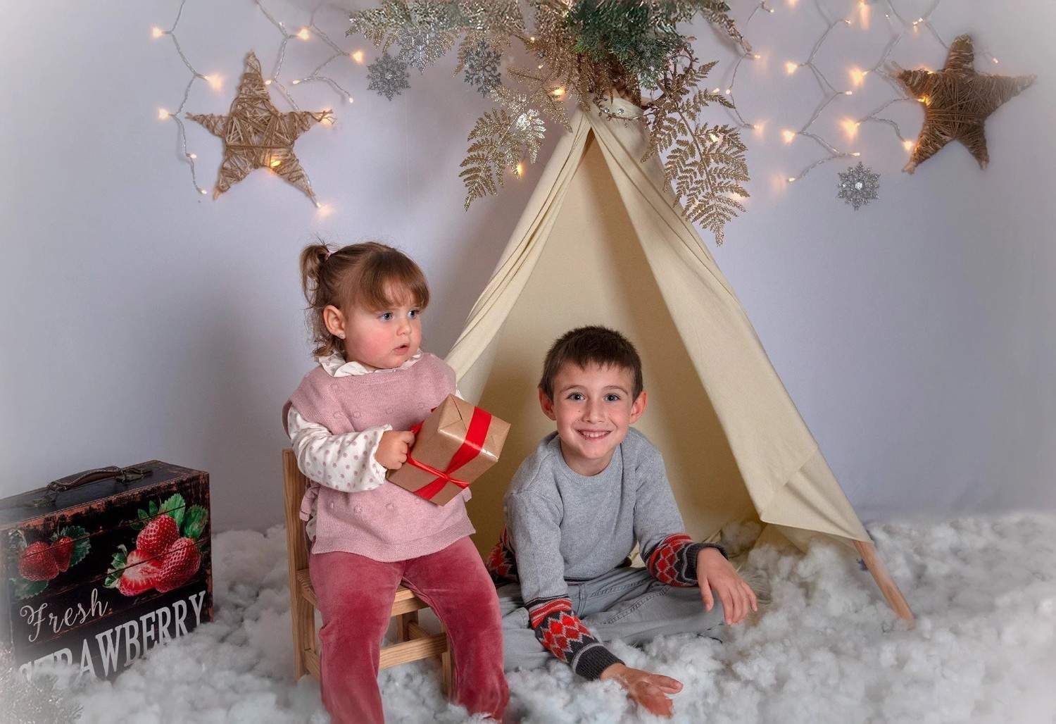 Dos niños sentados en una acogedora tienda de campaña navideña con luces de hadas, sosteniendo una caja de regalo y sonriendo.