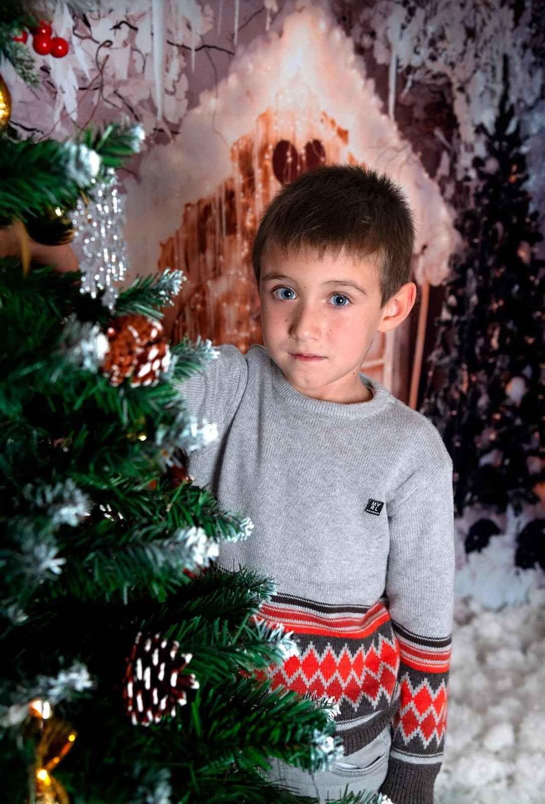 Niño con suéter gris de pie junto a un árbol de Navidad decorado en el interior de una casa.
