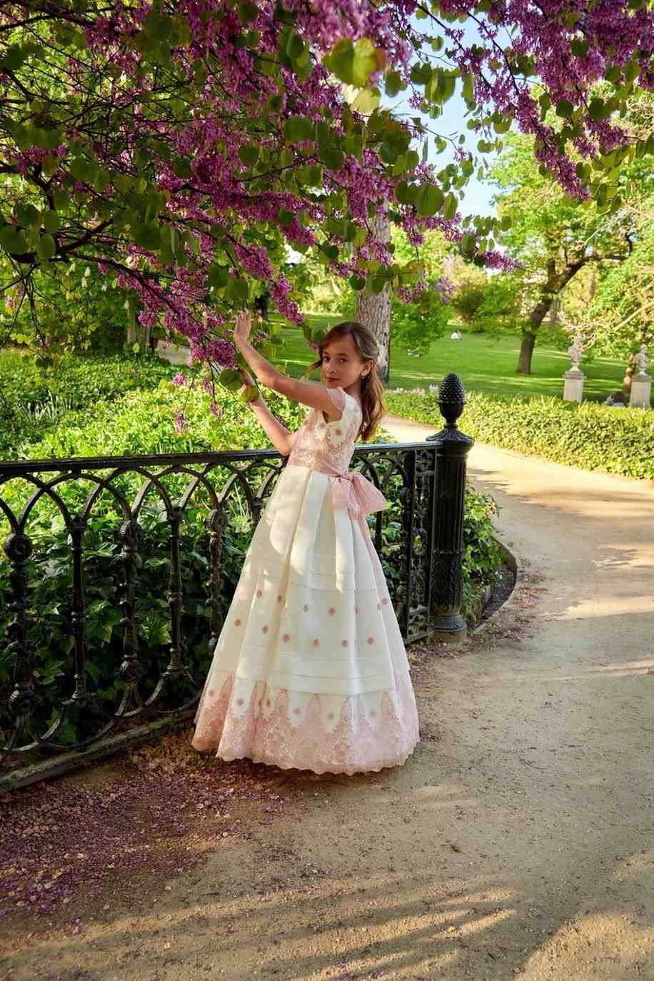 Mujer con vestido blanco posando bajo flores rosas en un sendero de jardín.