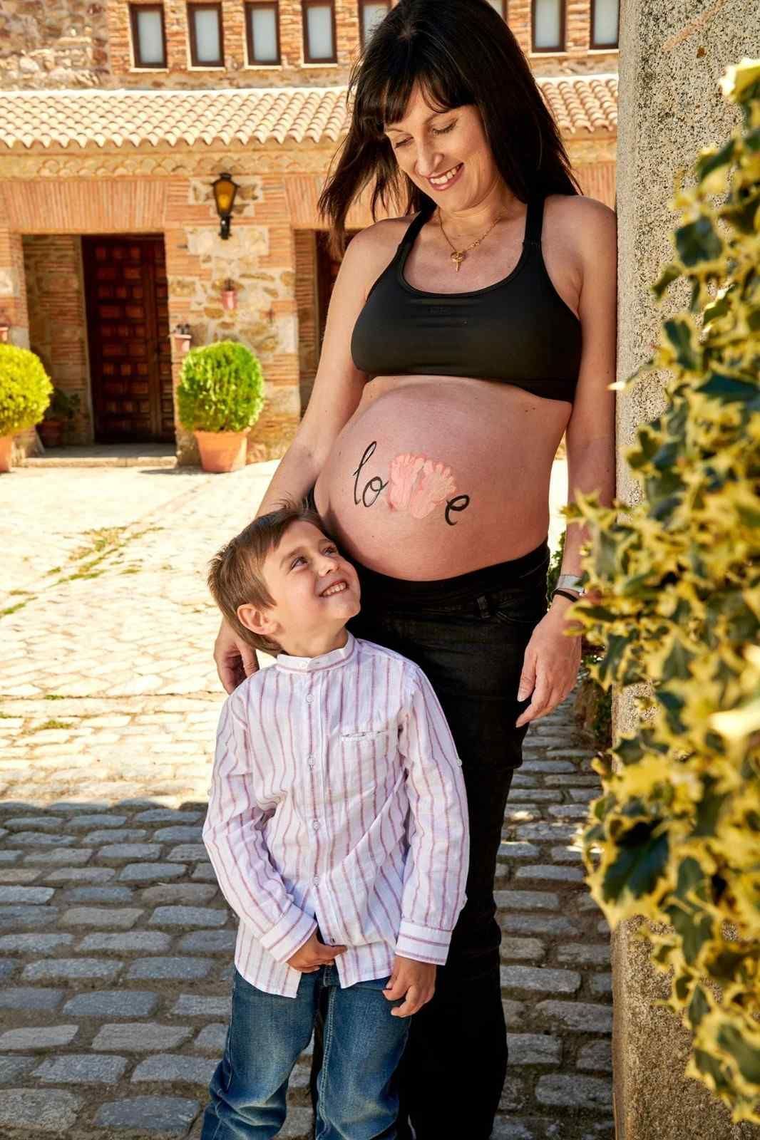 Una mujer embarazada y su hijo están de pie al aire libre en un patio soleado, ambos mirando hacia arriba y sonriendo.