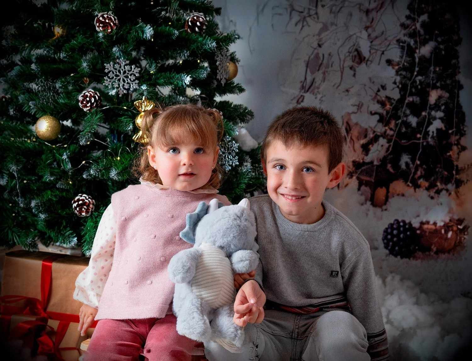 Dos niños están sentados junto a un árbol de Navidad decorado, sosteniendo un conejo de peluche gris, con regalos envueltos cerca.