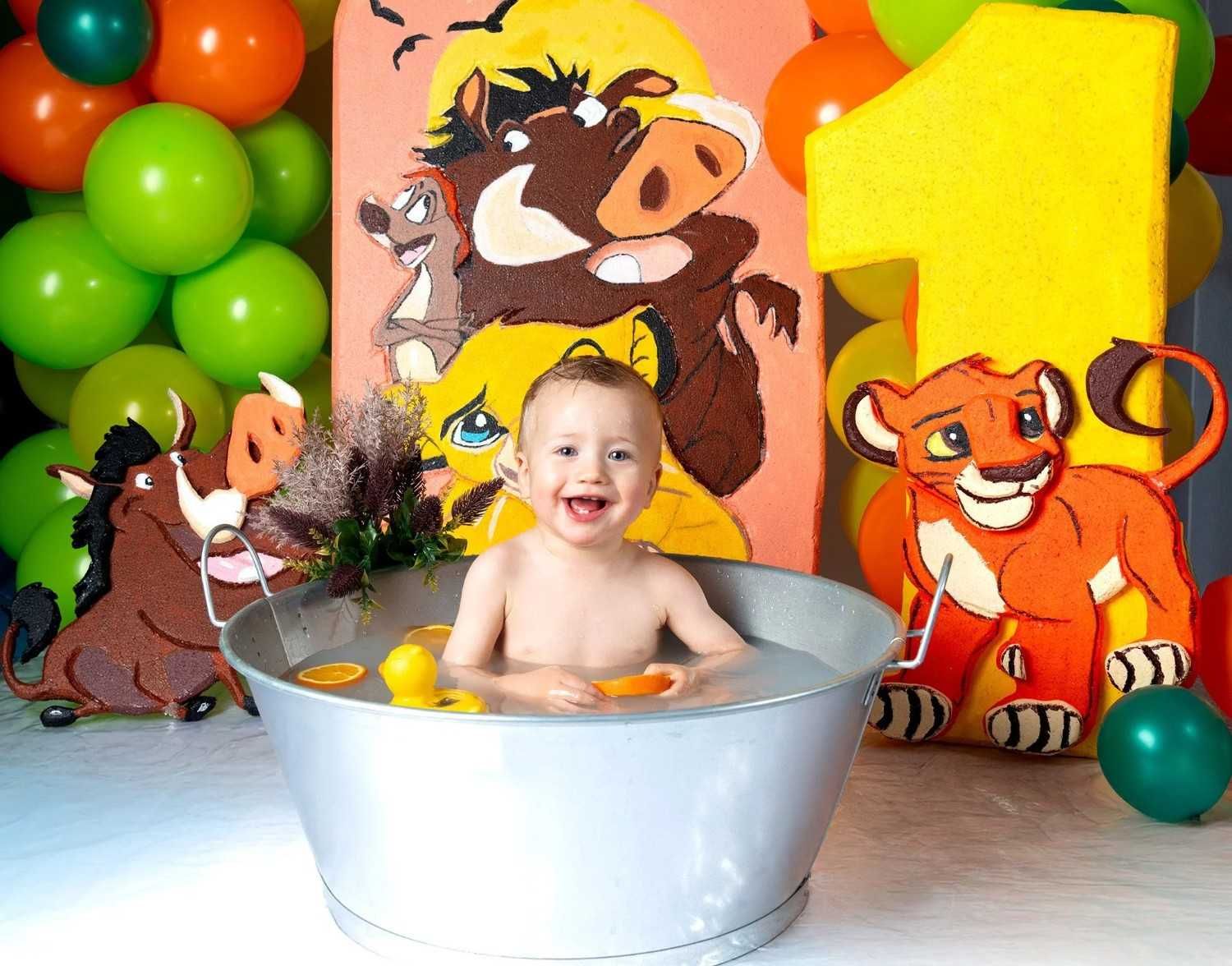 Bebé sonriente en una bañera plateada entre globos y decoraciones de fiesta con animales de dibujos animados.