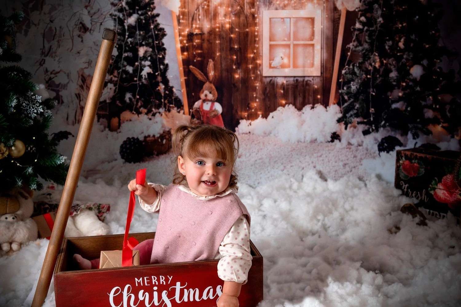 Niño en una caja roja de Navidad, sonriendo en un escenario navideño nevado con árboles y una ventana decorada.