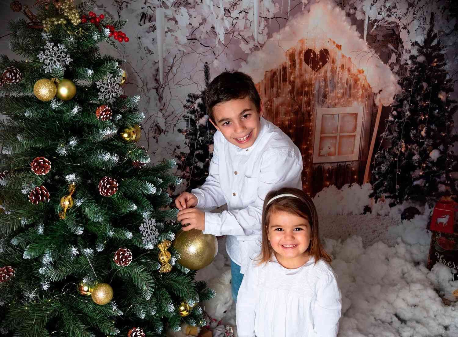 Dos niños sonriendo junto a un árbol de Navidad decorado en una escena navideña nevada.