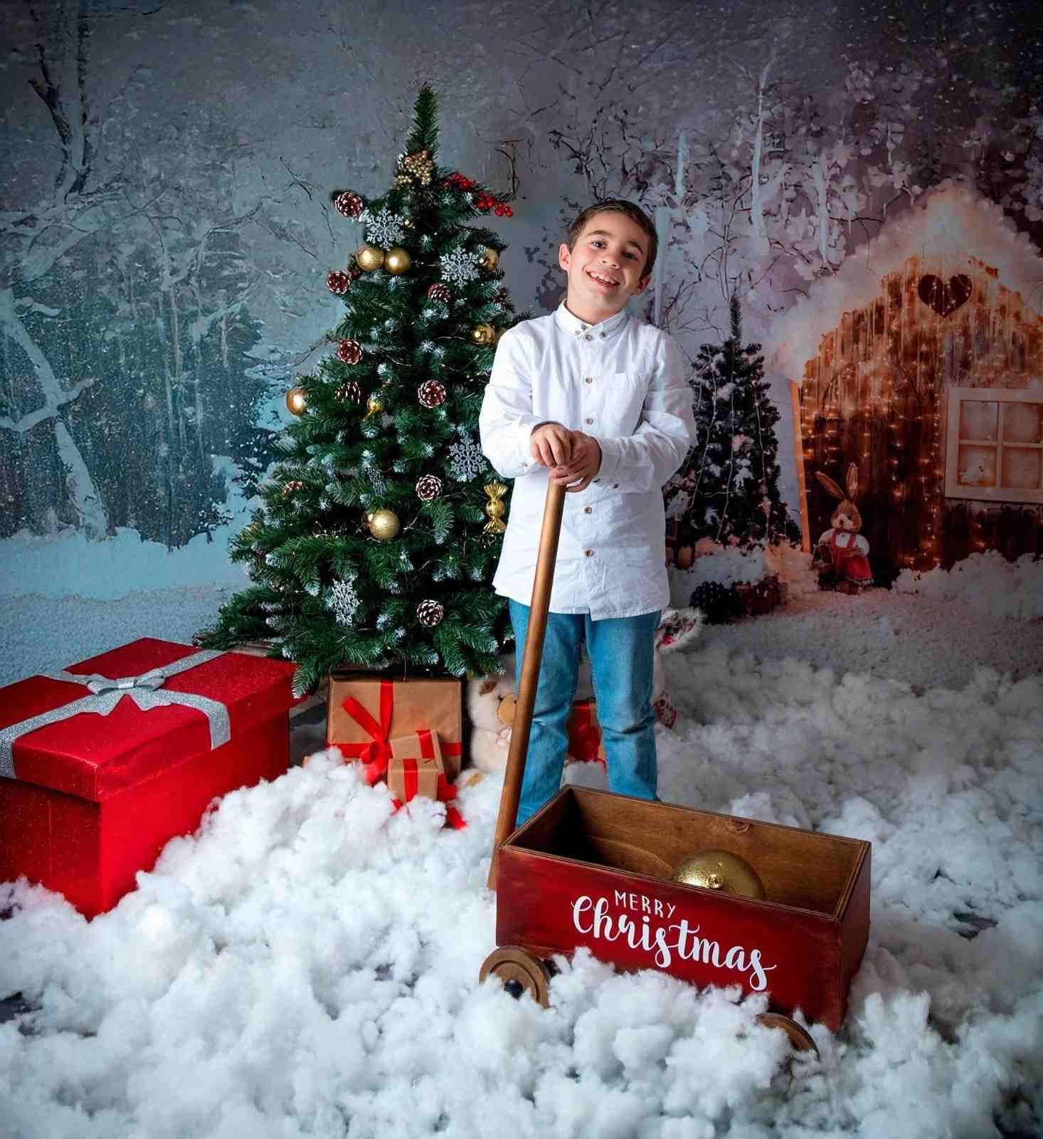 Un niño con camisa blanca está de pie junto a un árbol de Navidad con regalos y un carrito rojo navideño en una escena invernal nevada.