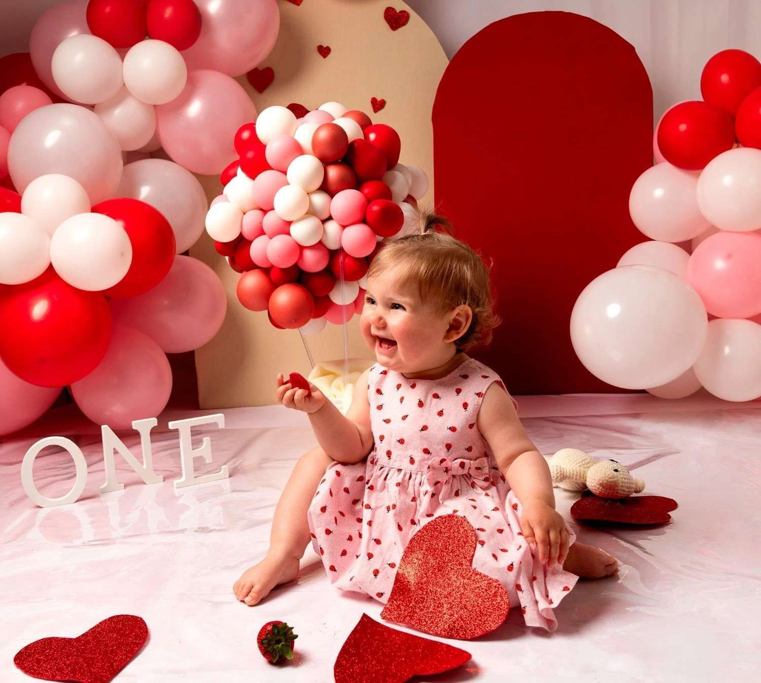Bebé con vestido rosa sentada entre globos rojos y blancos en forma de corazón para una sesión de fotos con temática de San Valentín.