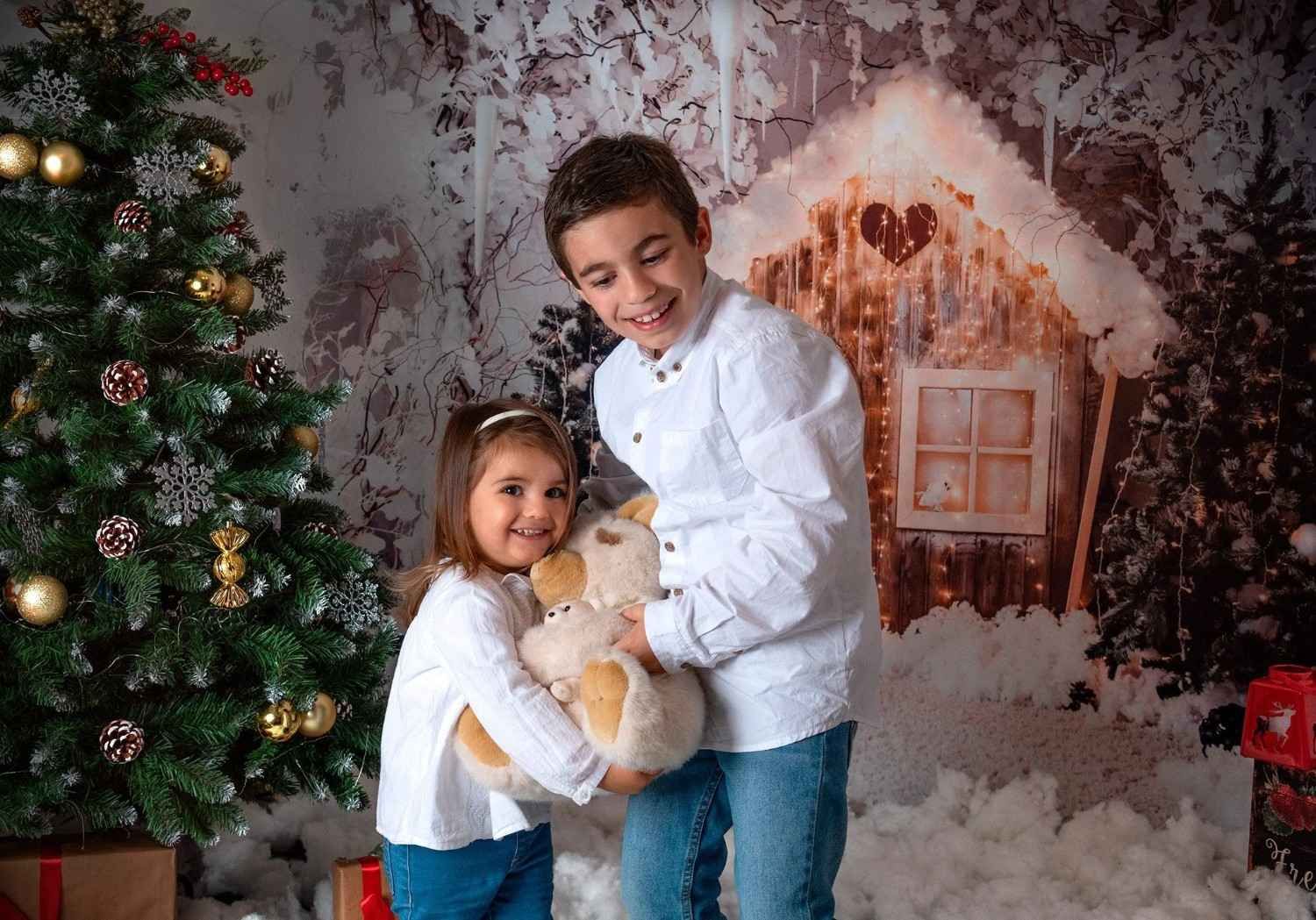 Dos niños con suéteres blancos abrazan un osito de peluche junto a un árbol de Navidad decorado y un fondo festivo.