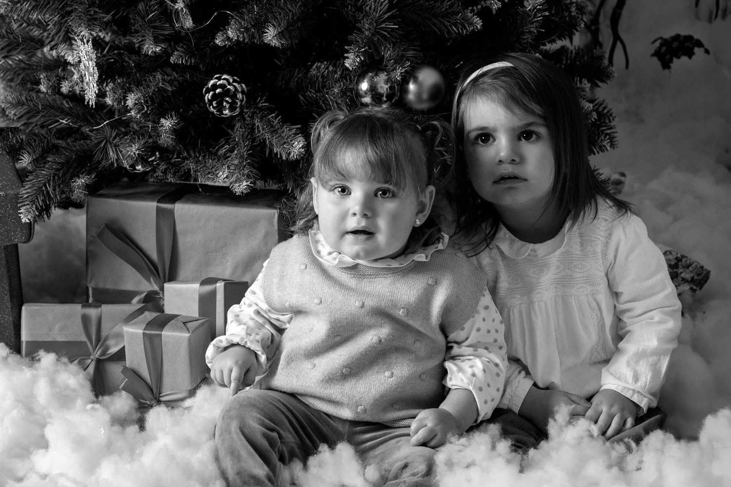 Dos niños sentados junto a regalos envueltos bajo un árbol de Navidad, rodeados de esponjosas decoraciones blancas.
