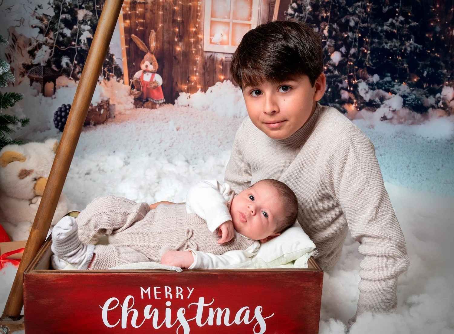 Dos niños vestidos de blanco en una escena navideña nevada, con un bebé acostado en una caja roja con la inscripción 