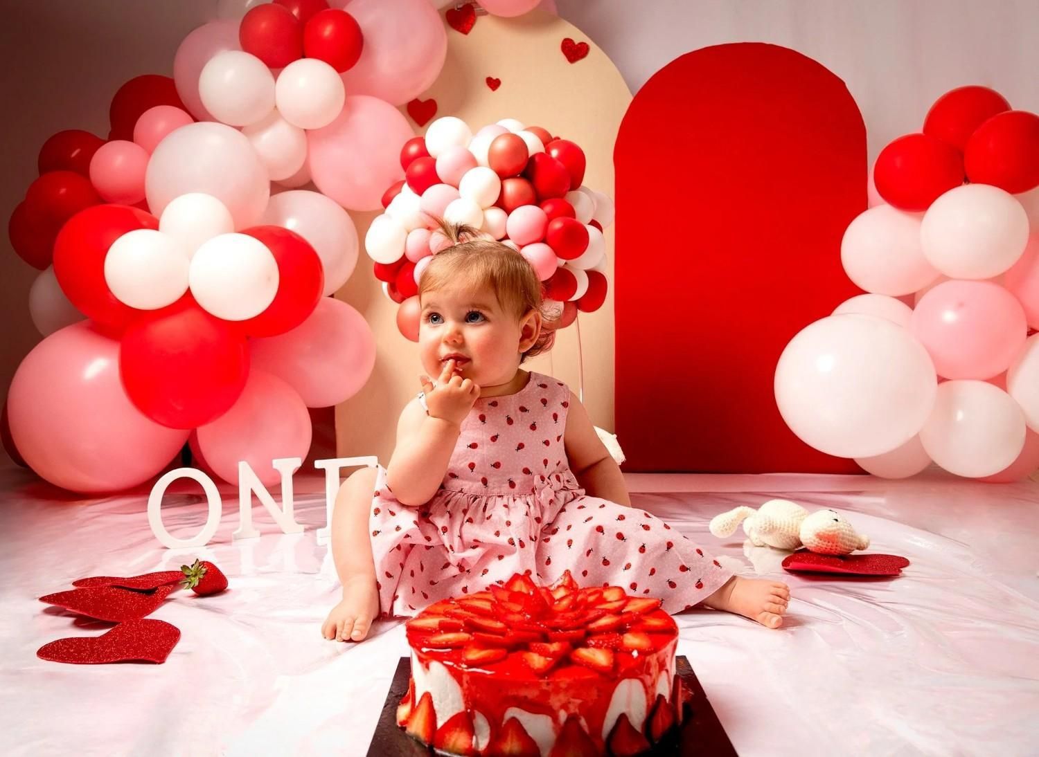 Niña con vestido rosa de lunares en una decoración de cumpleaños con globos rojos y rosas y un gran pastel rojo.