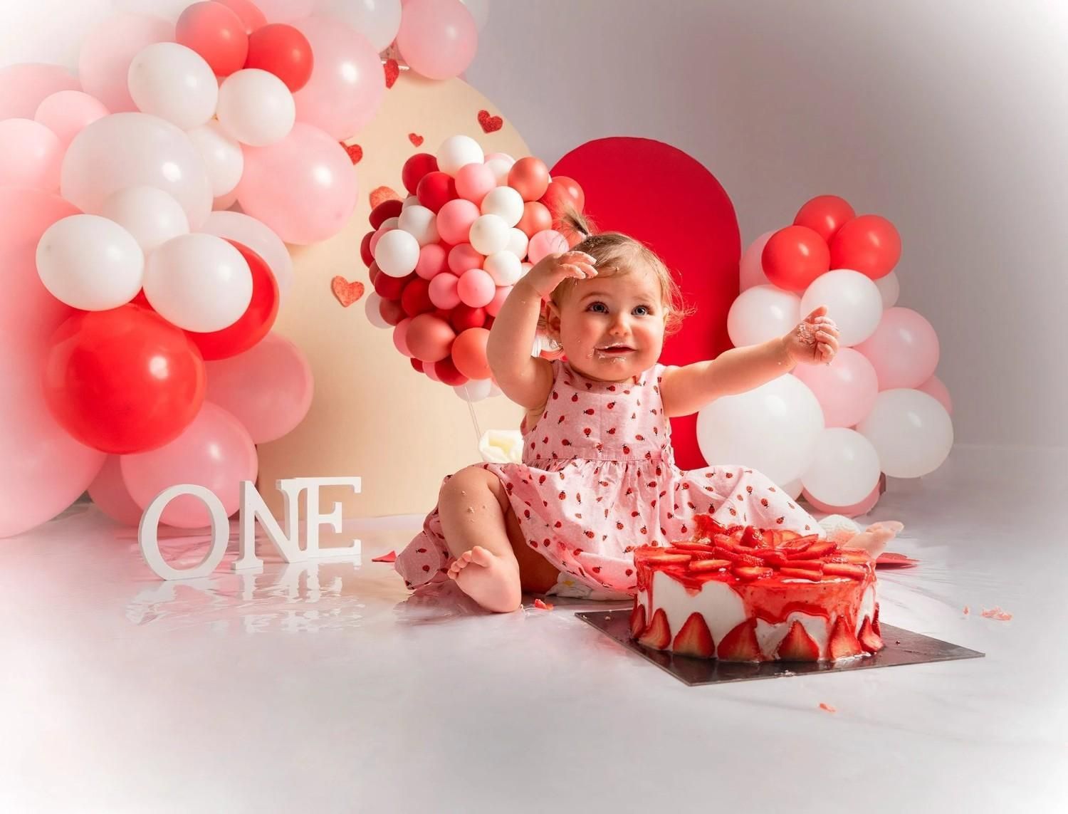 Bebé celebrando su primer cumpleaños entre globos rojos y blancos con una caja de regalo y un letrero que dice 