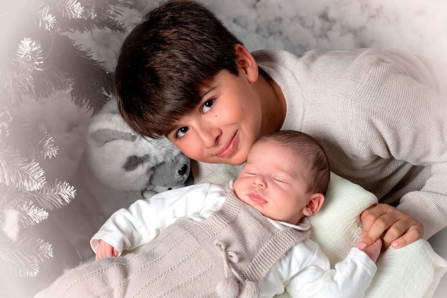 Dos niños acurrucados en una manta suave, uno sonriendo a la cámara mientras sostiene a un bebé dormido.