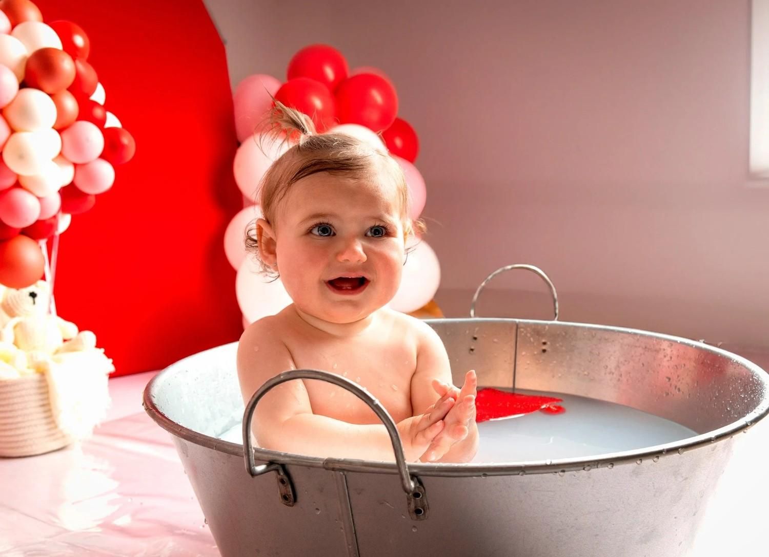 Bebé sonriente en una bañera plateada con globos rojos y blancos al fondo.