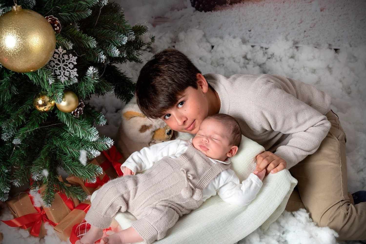 Un niño y un bebé yacen junto a un árbol de Navidad decorado, ambos envueltos en suaves mantas.