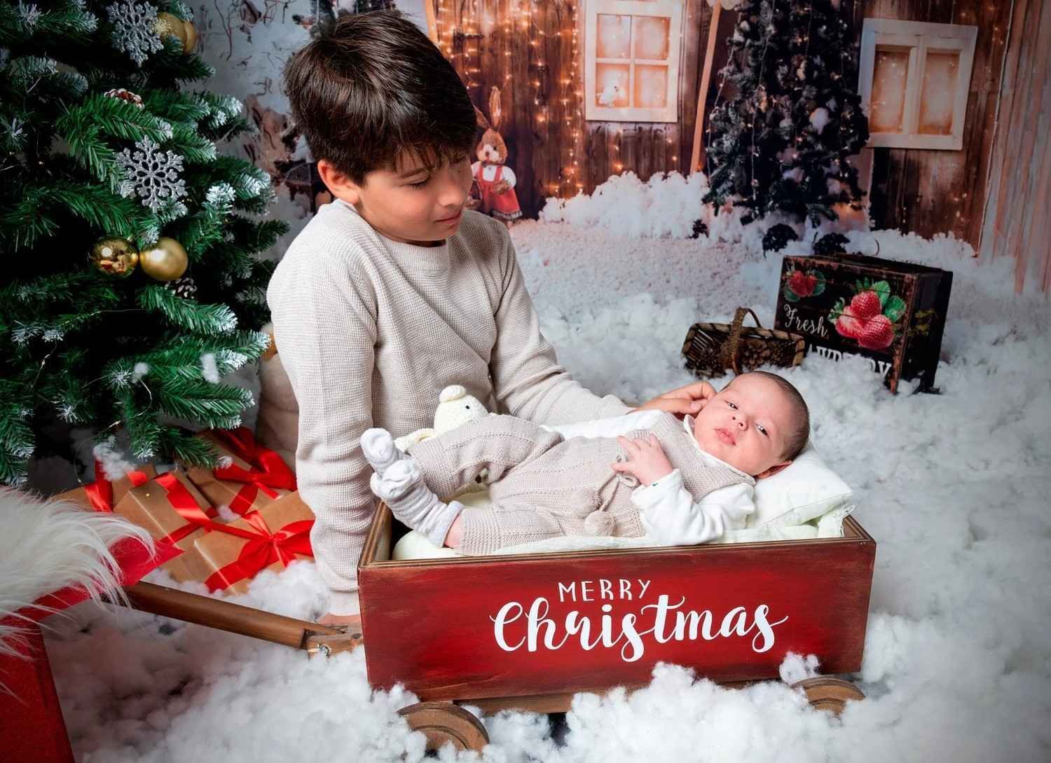 Un niño sostiene a un bebé dormido en una caja roja de Navidad junto a un árbol decorado y regalos.