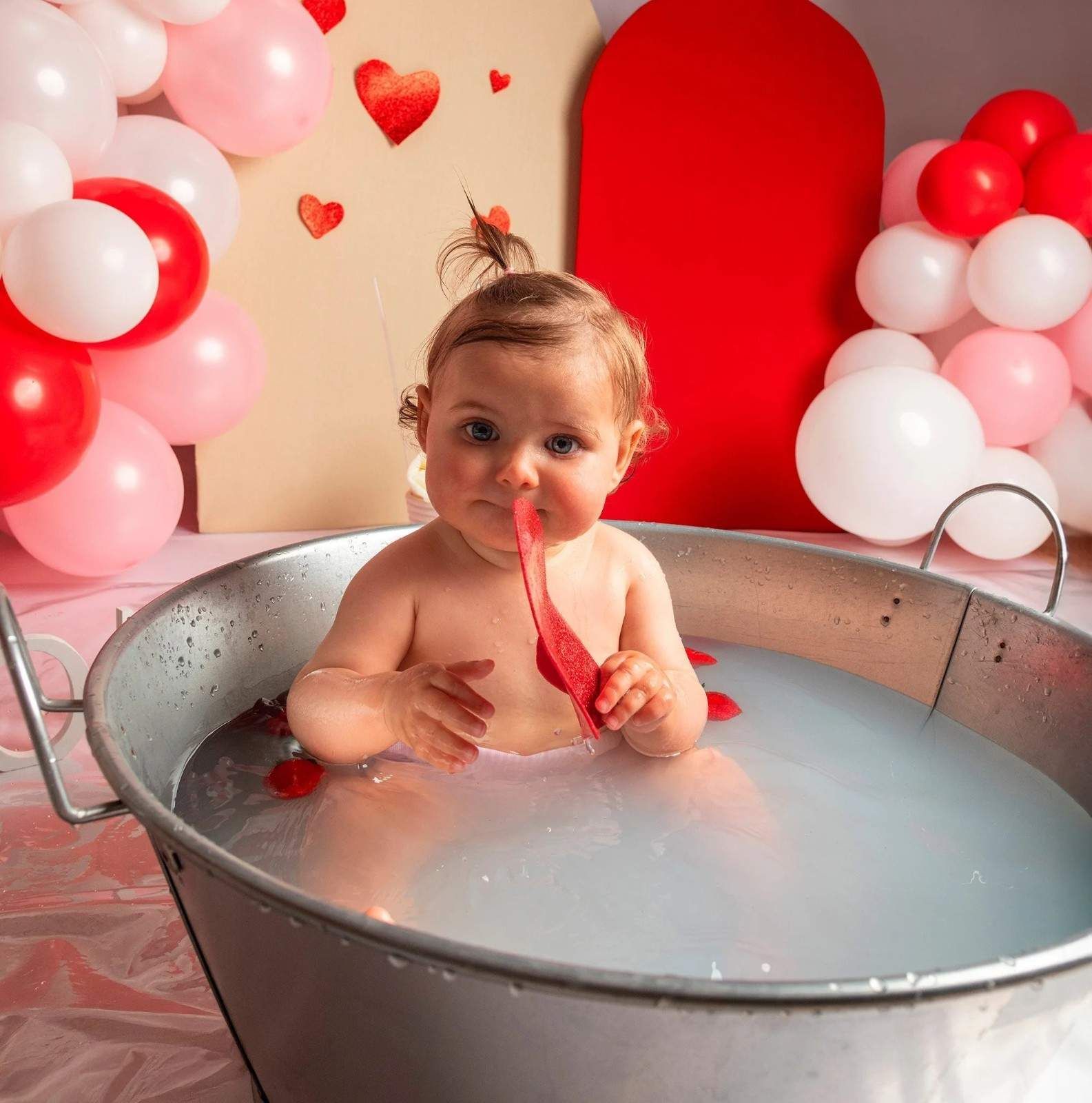 Bebé en una bañera de metal rodeado de globos rojos y blancos, sosteniendo una cinta roja en un ambiente festivo.
