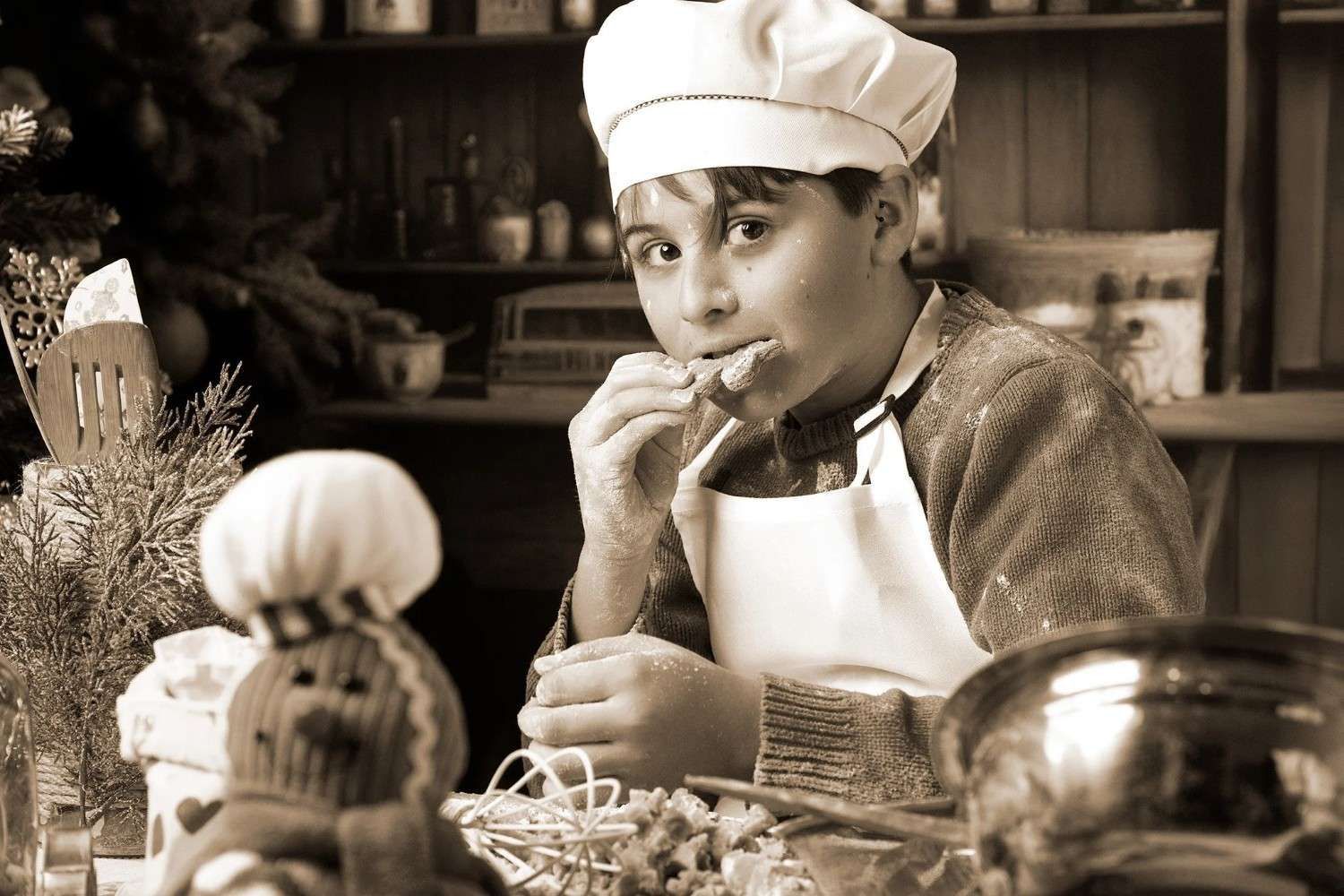 Joven chef con gorro y delantal blancos degustando comida en una cocina rústica con ingredientes y utensilios de cocina.