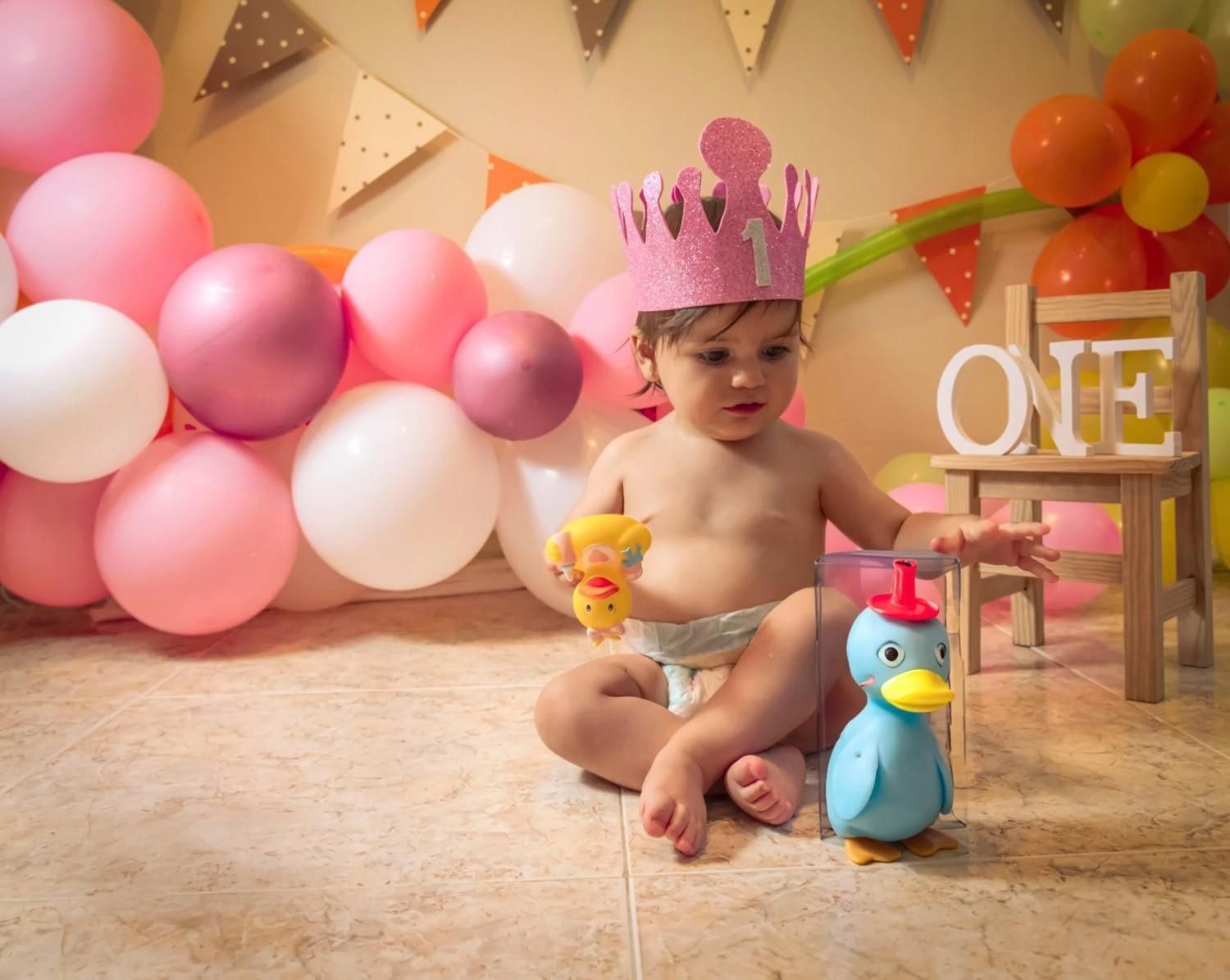 Una niña pequeña con una corona rosa entre globos de colores pastel, sosteniendo un patito de juguete en la decoración de una fiesta de cumpleaños.