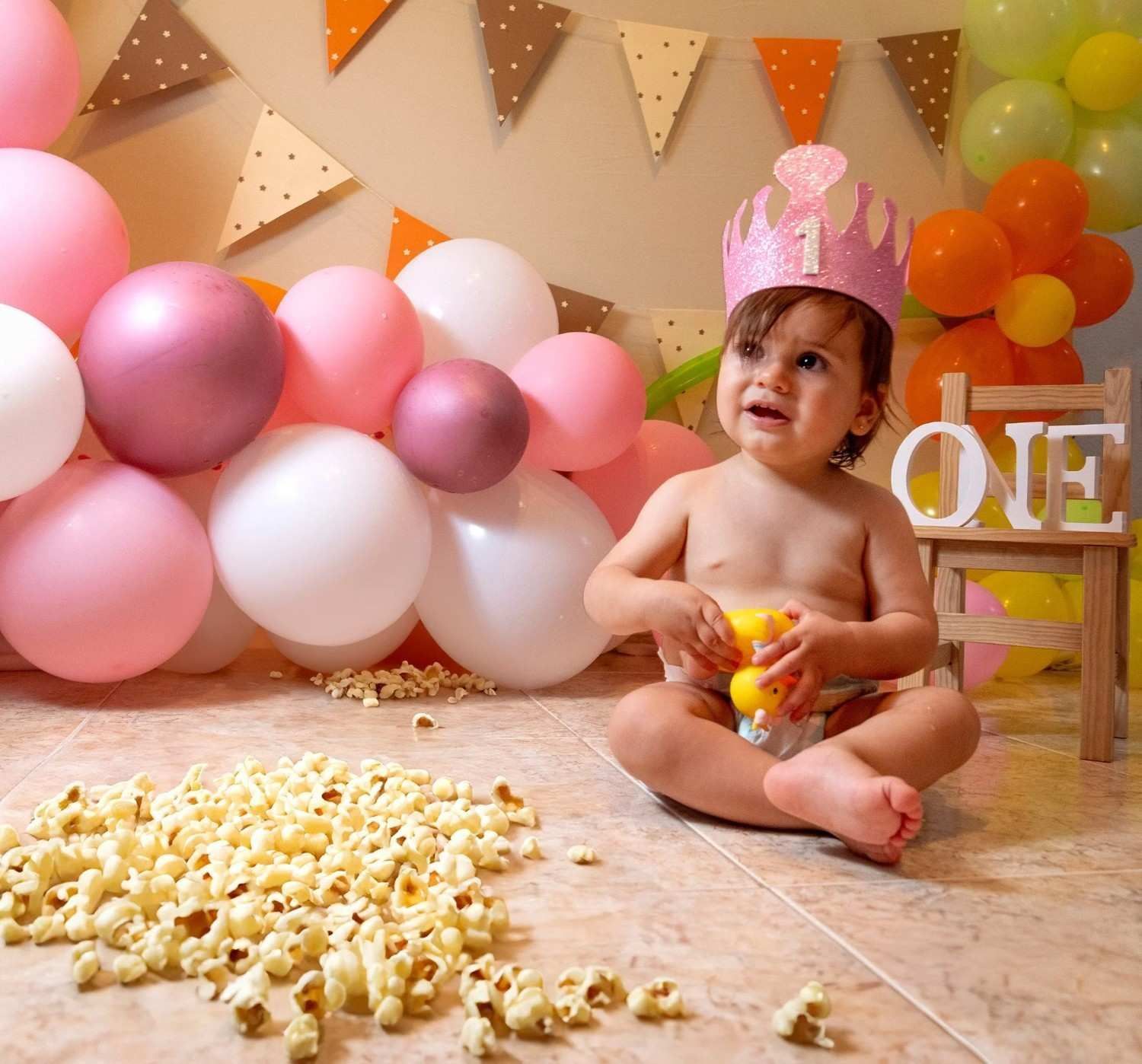 Bebé con gorro de fiesta rosa sentado entre globos y palomitas de maíz en una celebración de su primer cumpleaños.