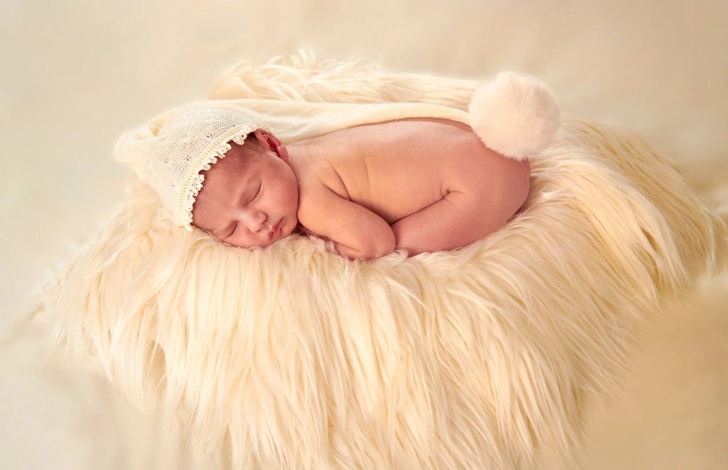Recién nacido durmiendo acurrucado sobre una suave manta color crema, con un gorrito de punto, en una luz cálida y suave.