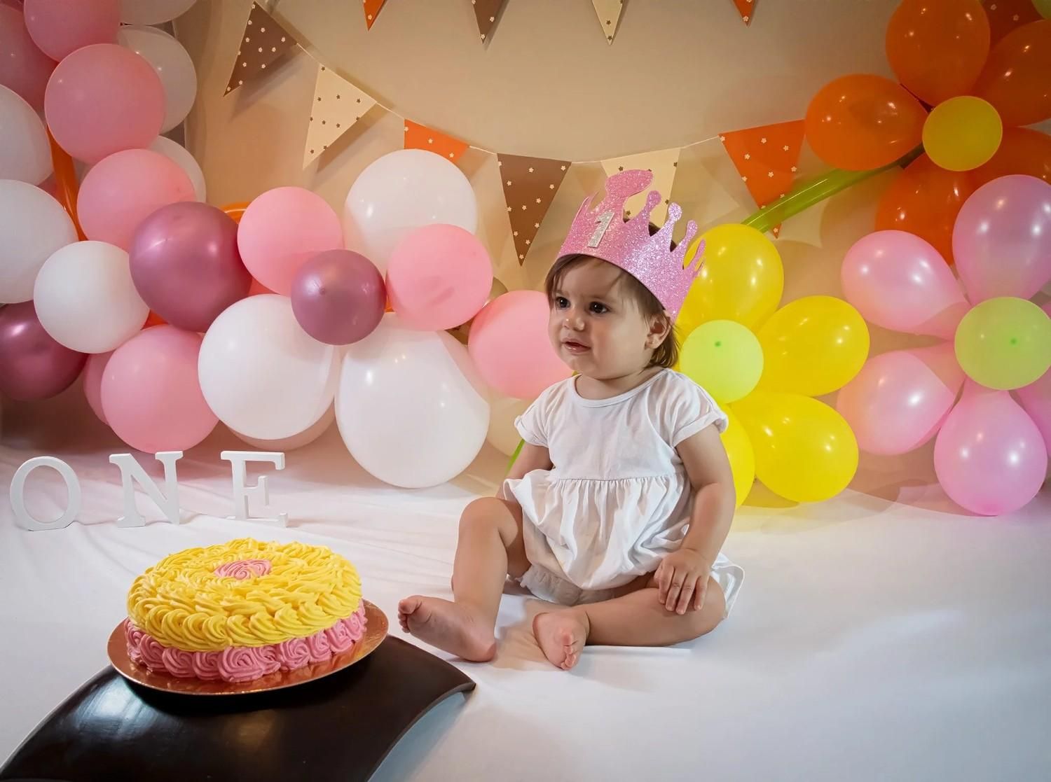 Un niño sentado junto a una colorida tarta de cumpleaños entre globos y guirnaldas de colores pastel.