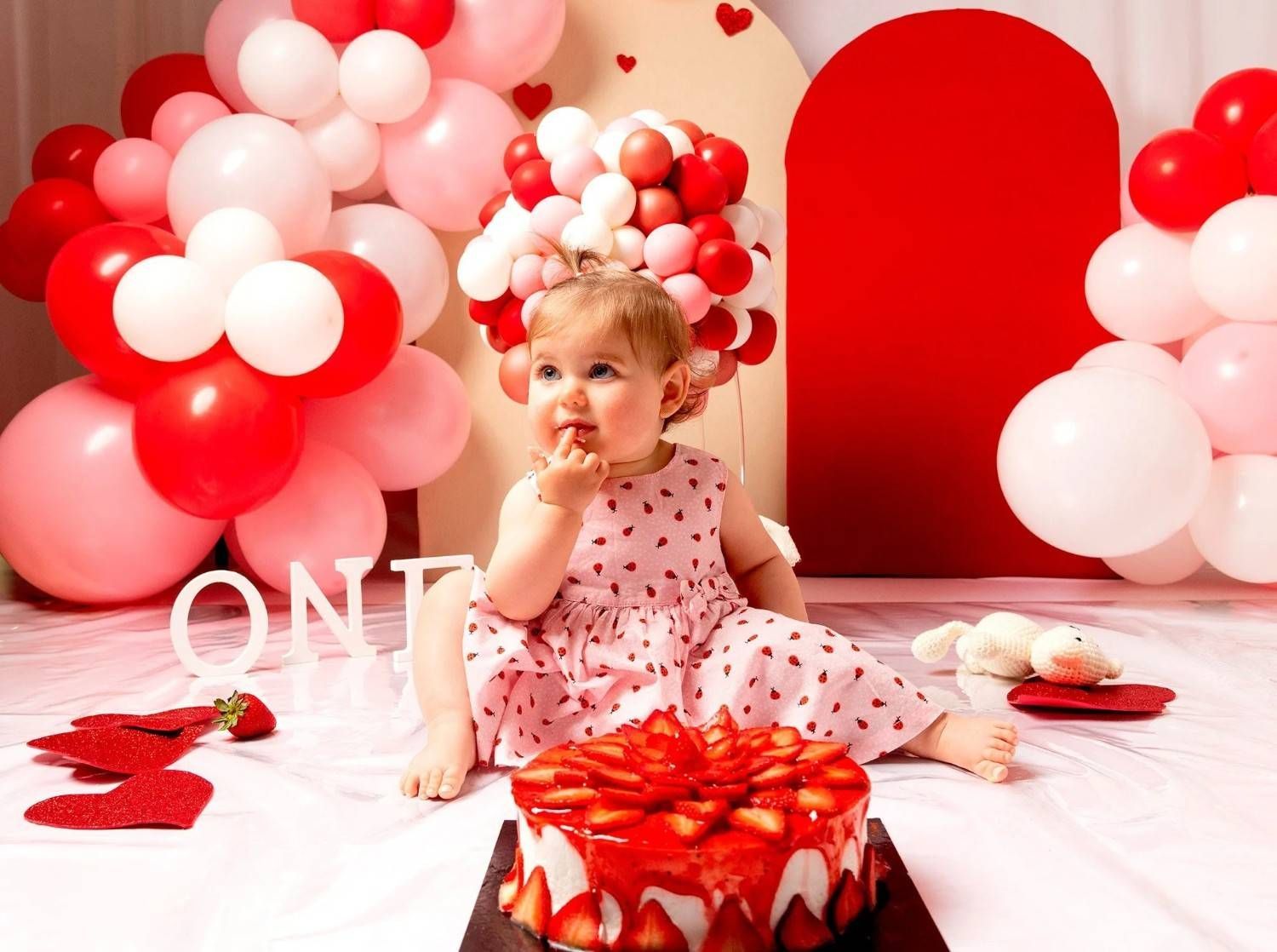 Niña pequeña con vestido rosa en una fiesta de primer cumpleaños roja y blanca con globos y pastel de fresa.