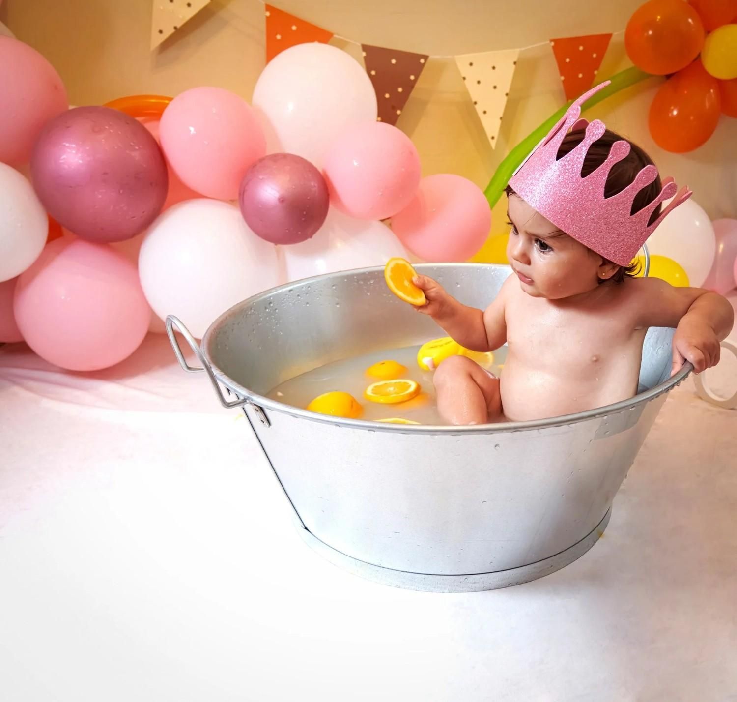 Bebé en una bañera plateada con globos y guirnaldas rosas, con una corona rosa y sosteniendo juguetes de baño amarillos.