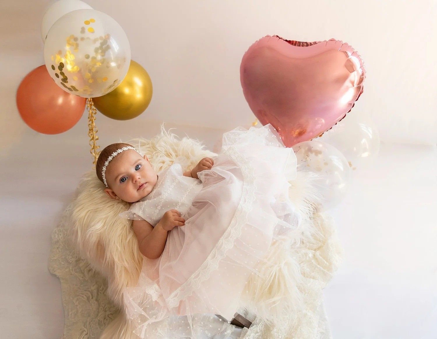 Bebé con un vestido de tul blanco recostada junto a globos en forma de corazón dorados, color melocotón y rosa sobre un fondo blanco.