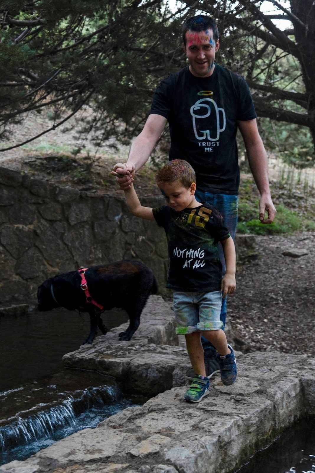 Un adulto y un niño pasean a un perro negro por un sendero de piedra junto a un arroyo.