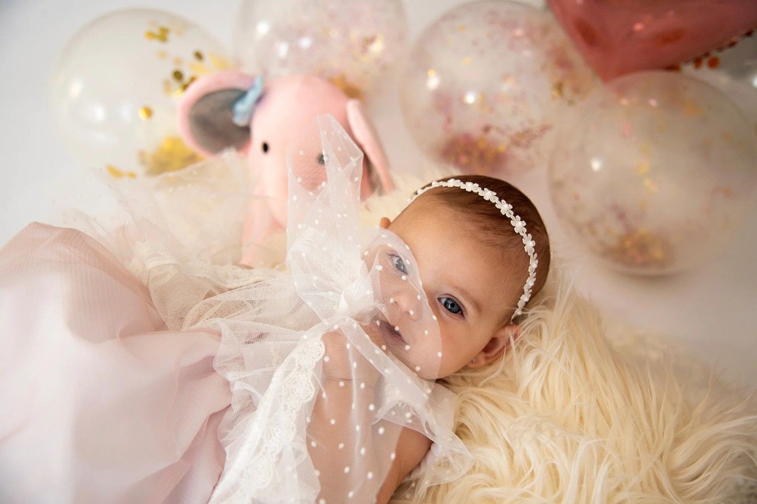Bebé tumbado sobre una manta mullida con globos de colores pastel y un vestido de tul blanco.