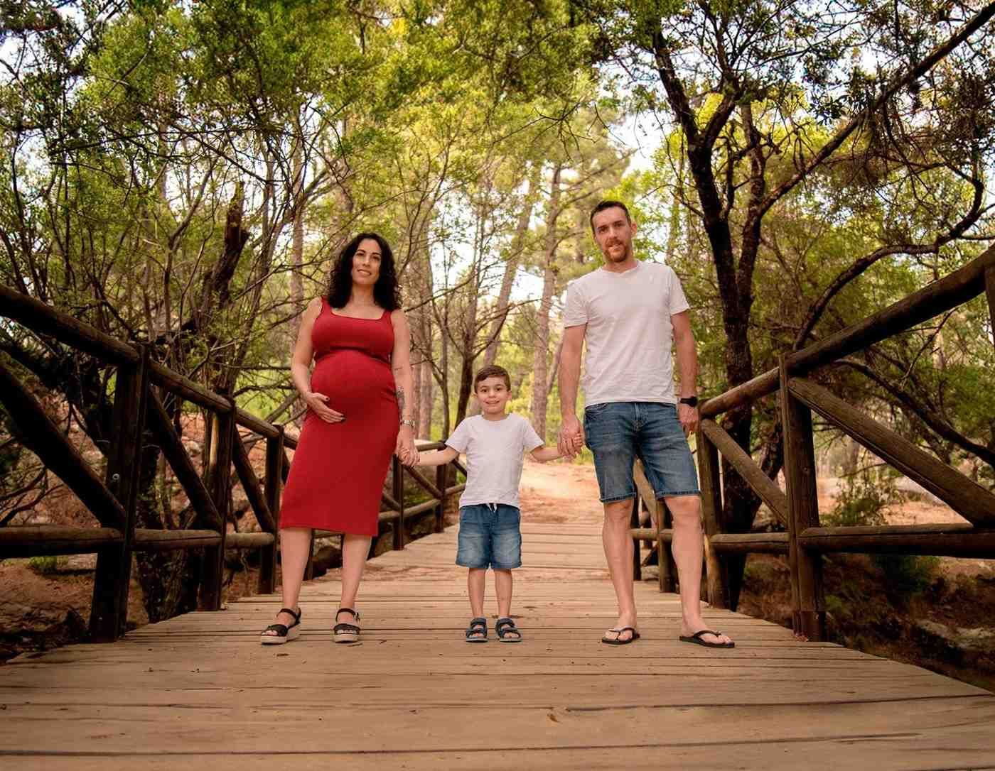 Tres personas caminan de la mano sobre un puente de madera en un parque arbolado, bajo la sombra de los árboles.